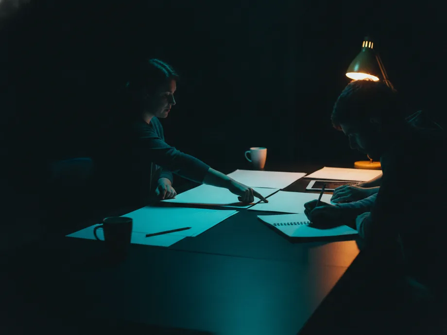 Two people working across a long dark table at night, one pointing at a printed plan while the other writes in a notebook under a warm lamp.