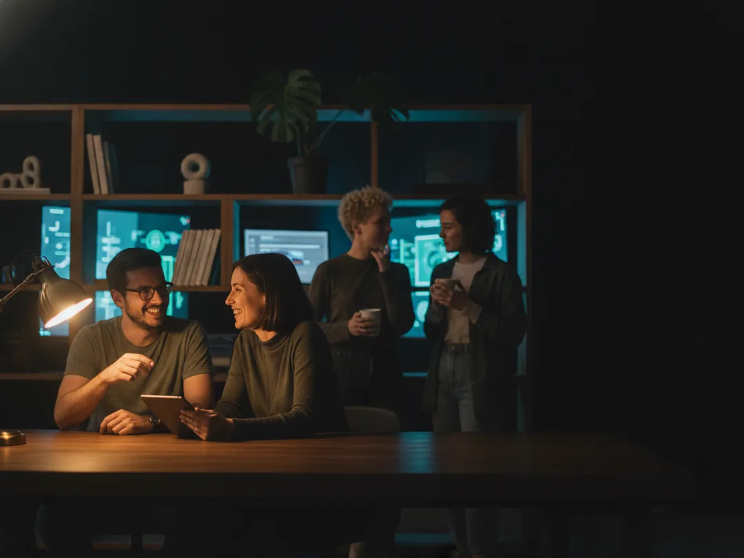 Two colleagues pairing at a desk in the studio while two teammates talk over coffee behind them.