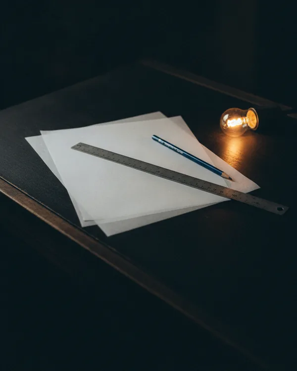 Blank sheets of tracing paper layered on a dark drafting table under a metal ruler and a pencil.