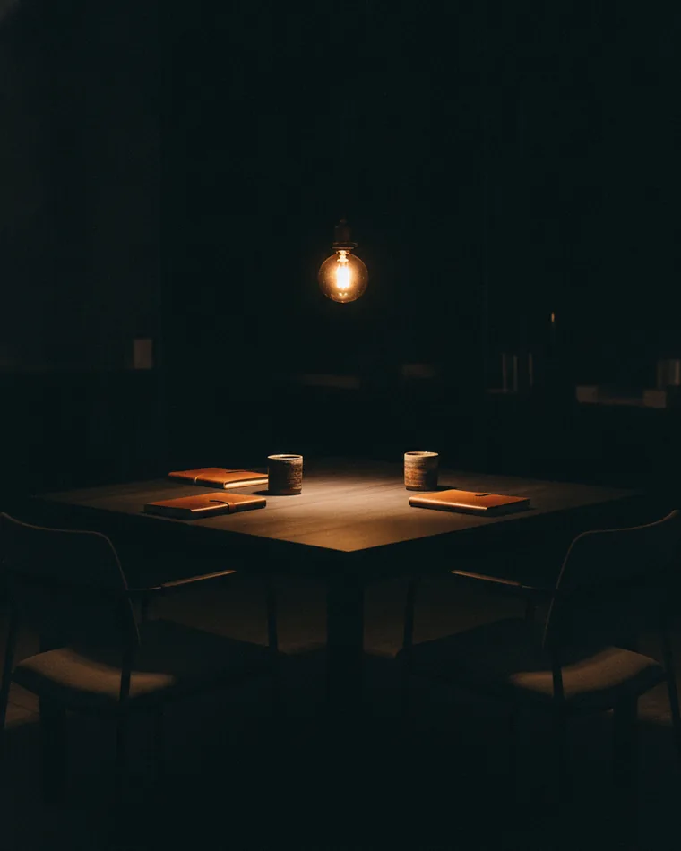 Two pulled-out chairs at one long dark table under a single pendant lamp, a notebook and a cup at each place.