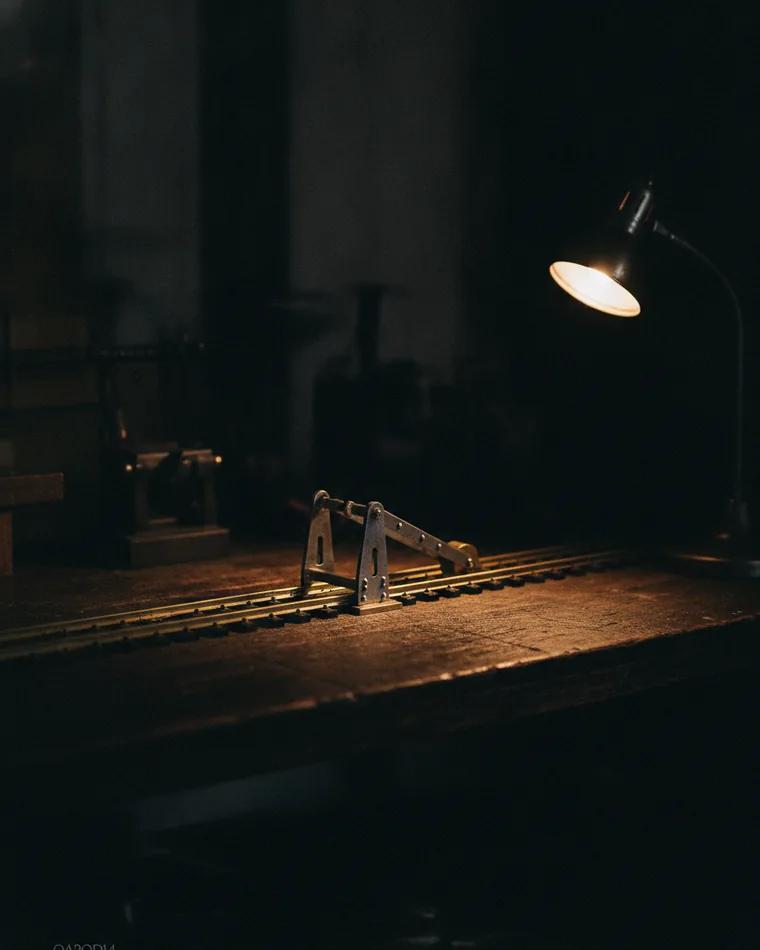 A brass rail track on a dark bench interrupted by a small hinged gate, held open by a weighted lever.