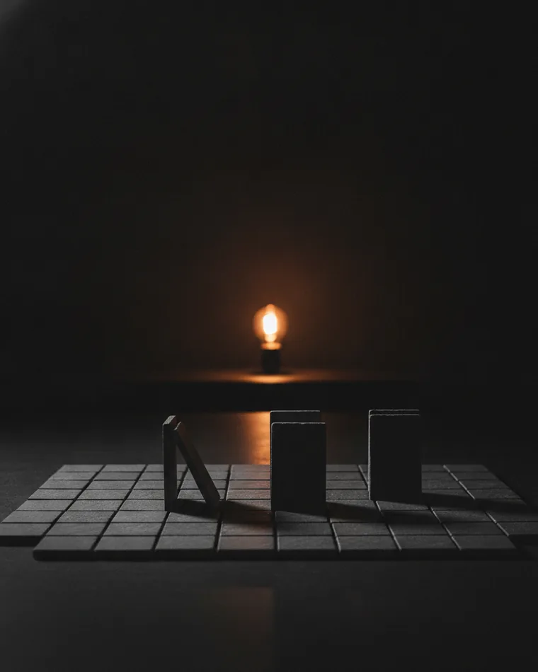 A grid of small slate tiles laid out across a dark table with three tiles standing on edge, lit by a single warm bulb behind.
