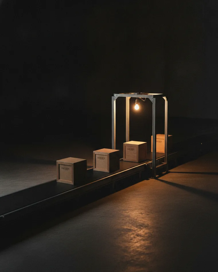 A short conveyor belt on a dark floor carrying three identical crates towards a lit gate.