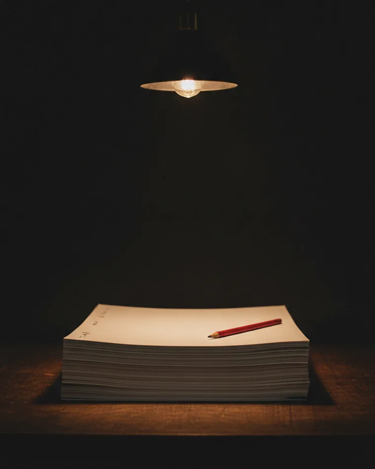 A stack of loose printed sheets squared on a dark table with a marked-up top sheet and a red pencil.