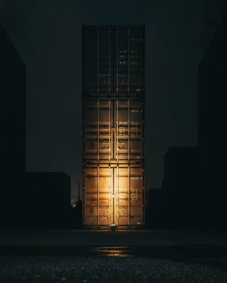 Identical steel shipping containers stacked in a dark yard with one bay lit from below.