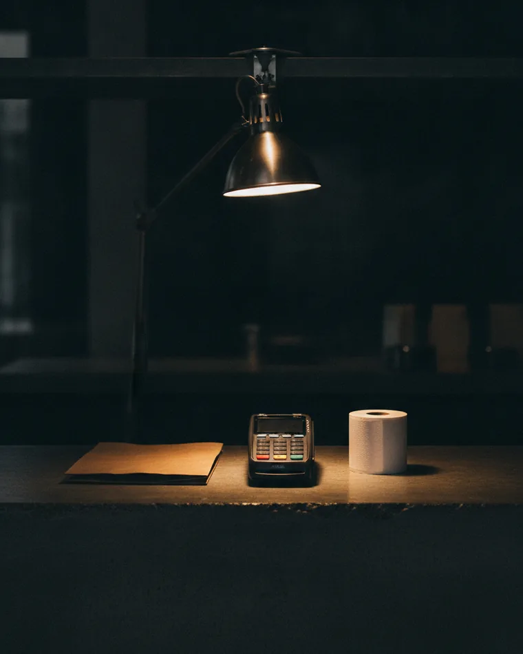 A dark shop counter with a card terminal, a paper roll and a folded bag under one lamp.