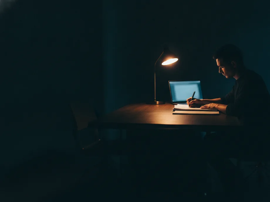 A writer marking up printed pages at a dark desk at night, lit by a single warm lamp beside an open laptop.