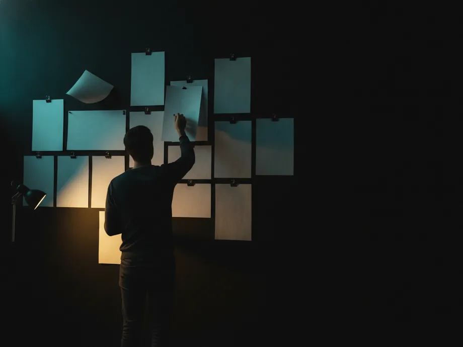 A person seen from behind pinning a blank sheet onto a dark studio wall already covered with a grid of blank pinned pages.