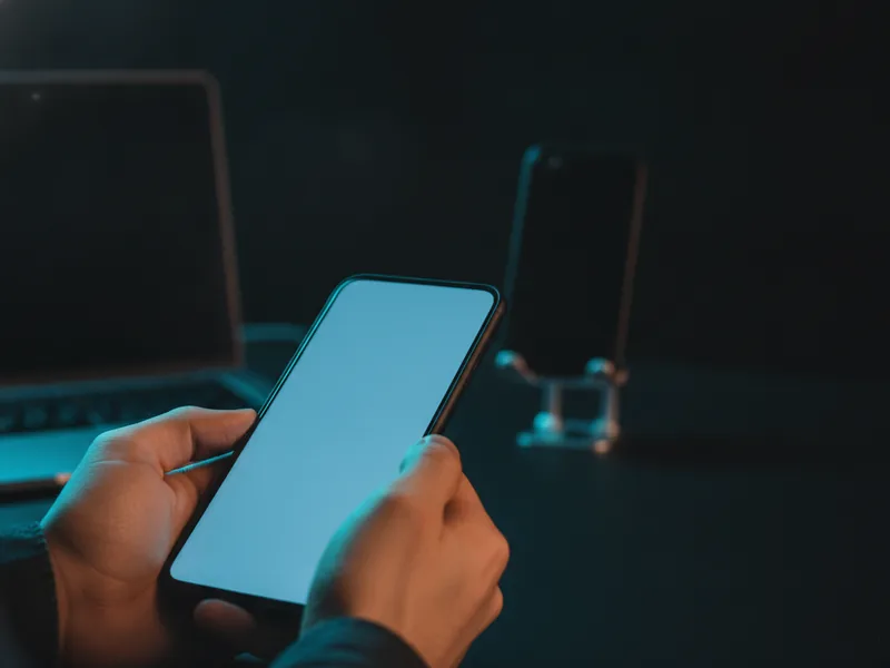 Two hands holding a phone with a soft blue screen, a laptop and a second device behind it.
