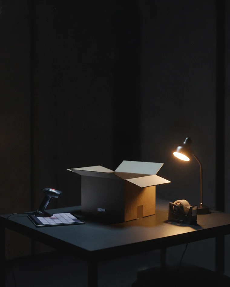 An open parcel, a barcode scanner and a tablet on a dark packing counter under one warm lamp.