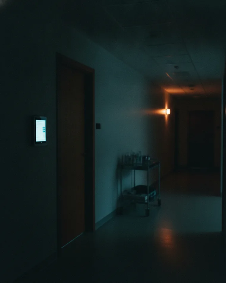 A quiet clinic corridor at night, a wall tablet station glowing beside a closed consulting-room door.