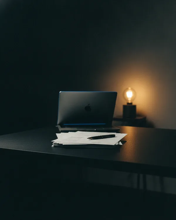 A squared stack of blank printed sheets on a black desk, a laptop edge-lit behind.