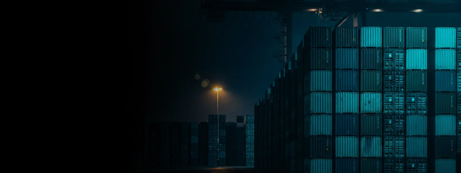 Stacked shipping containers in a night terminal, floodlit in a clean grid under a gantry crane.