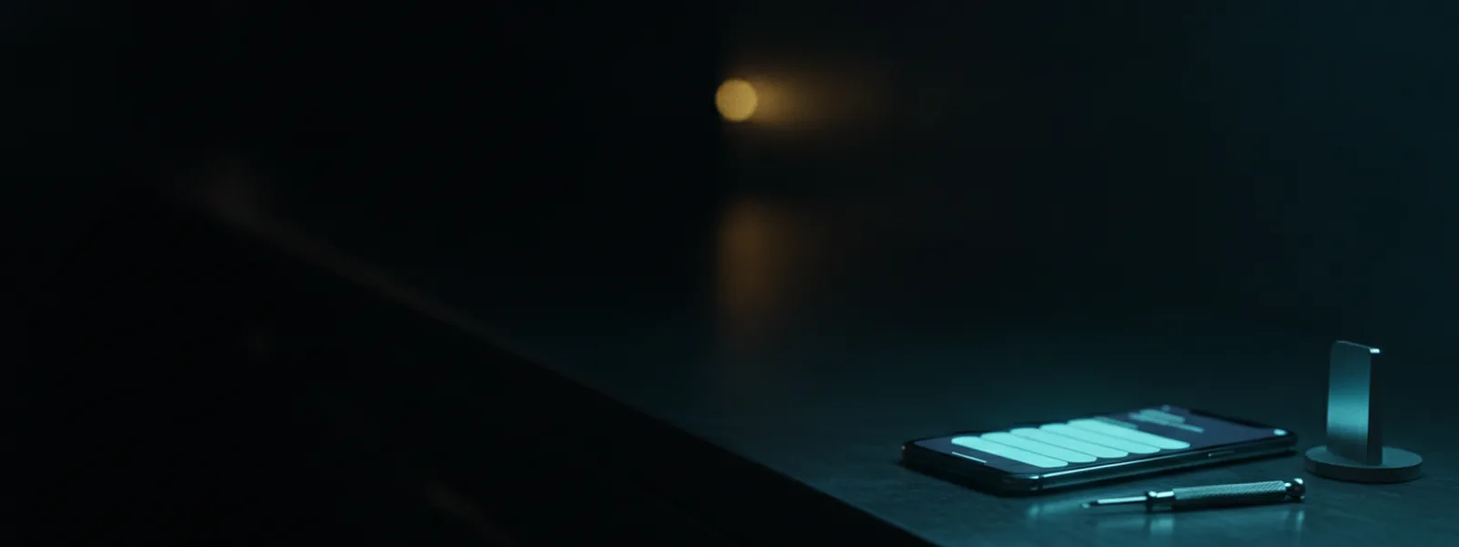 A phone face up on a dark metal bench beside a precision screwdriver, its screen glowing softly blue.