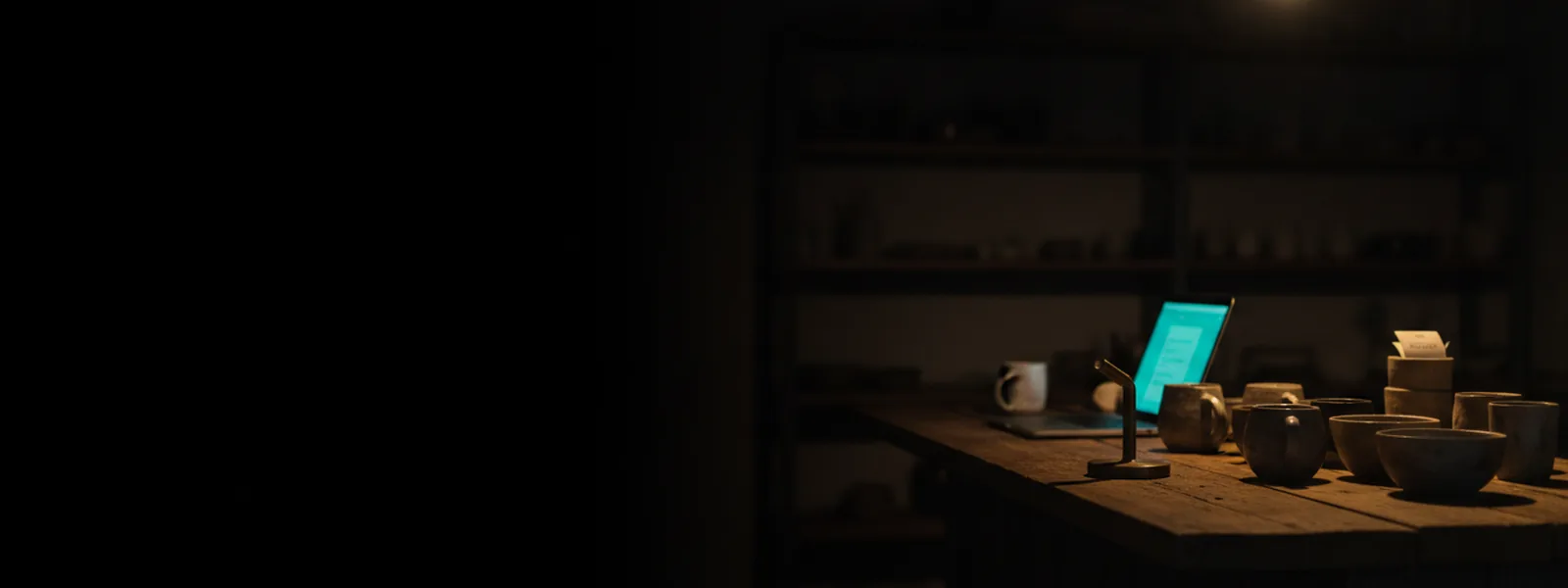 A small shop counter after closing, a laptop open beside a few plain ceramic products under a pendant lamp.
