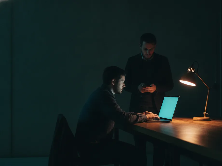 Two engineers at a long workbench at night, one typing on a laptop while the other checks a phone under a warm desk lamp.